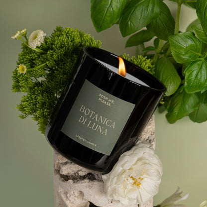 Lit Botanica di Luna candle among basil leaves and white flowers on a green textured surface