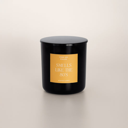 Smells Like The 80s black glass candle with golden yellow label by POSH ME, PLEASE., lid on, light background