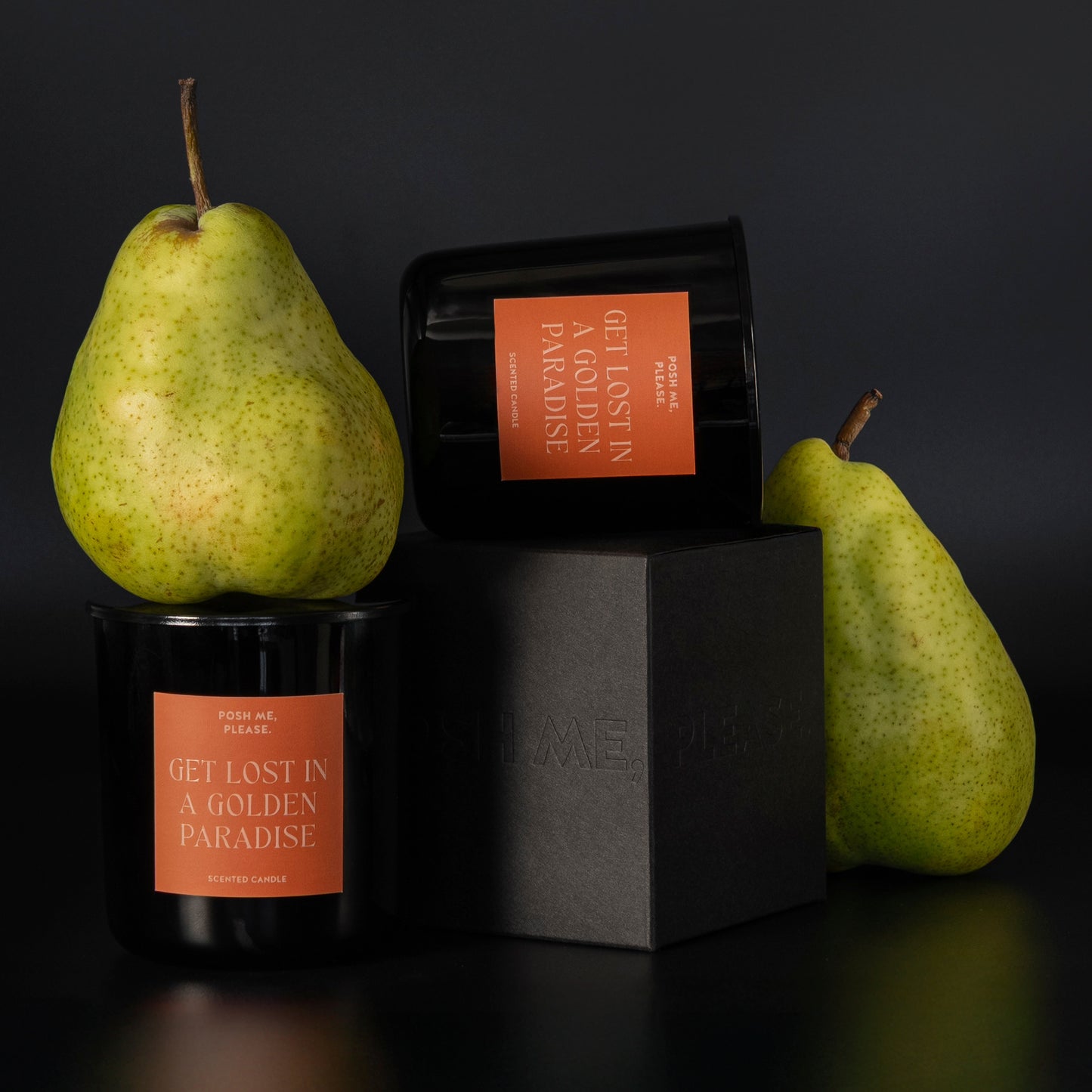 Two Get Lost in a Golden Paradise candles stacked on black blocks beside fresh green pears, dark background