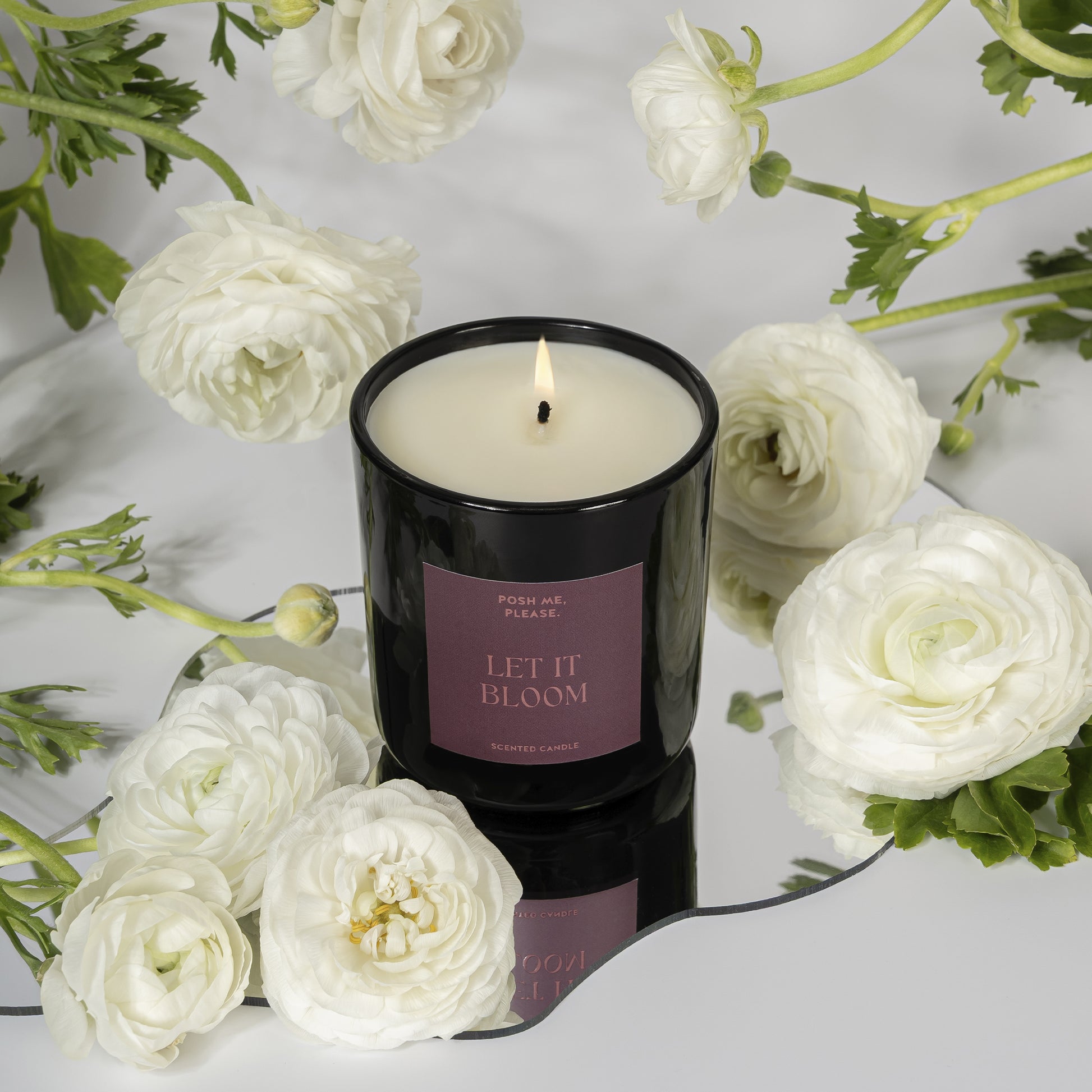Lit Let It Bloom candle surrounded by white roses and ranunculus flowers on a light grey surface